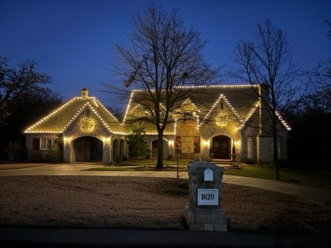 Stone home in warm white lights with lit wreaths and a bare tree out front